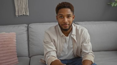 Modern bir oturma odasında oturan yakışıklı genç adam rahat bir tavırla gri koltukta rahat pembe yastıklı.