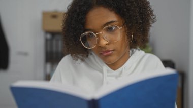 Güzel, kıvırcık saçlı ve gözlüklü Afro-Amerikalı bir kadın sessiz bir ofis ortamında kitap okuyor..