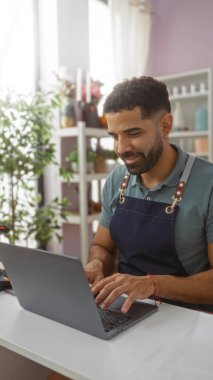 Ev dekorasyonu mağazasında dizüstü bilgisayarda çalışan genç İspanyol bir adam. Arka planda bitkiler ve raflar var.