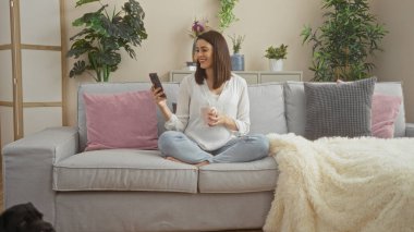 Genç, ispanyol bir kadın evdeki rahat bir kanepede dinleniyor, telefonuna gülümsüyor ve modern bir oturma odasında bitki ve yastıklı bir fincan tutuyor..