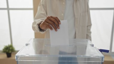 An african american man casting a vote by placing a ballot into a transparent box during an election held indoors. clipart