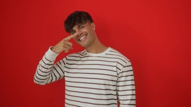 Yakışıklı İspanyol bir adam çizgili bir gömlek giyiyor, gülümsüyor ve yüzüne bakıyor, canlı kırmızı bir arka plana karşı duruyor..