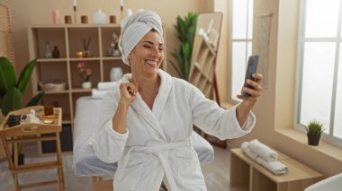 Güzellik merkezinde kafasında havluyla ve beyaz bir bornozla, sakin ve zarif bir iç mekanla selfie çeken bir kadın rahatlatıcı..