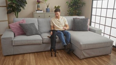 Genç, İspanyol bir adam, rahat bir oturma odasında, arka planda yastıklar ve bitkilerle süslenmiş rahat, gri bir kanepenin olduğu daksund 'uyla oynuyor..