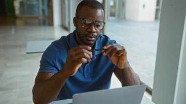 Mavi tişörtlü genç bir adam ofis içinde dizüstü bilgisayarla çalışıyor ve kalem üzerinde çalışıyor.