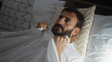 Modern bir yatak odasında yatağında sakallı, beyaz çarşafları yakın tutarken düşünceli görünen, evde yakalanmış İspanyol bir adam..