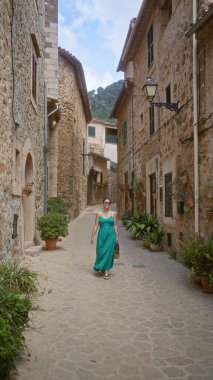 Valldemossa, Mallorca, İspanya 'da, taş binalar ve yemyeşil bitkilerle çevrili yeşil bir elbise ve güneş gözlüğü takmış, resim gibi dar bir sokakta yürüyen bir kadın..
