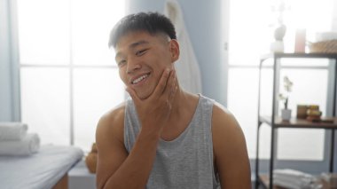 Kaplıca odasında gülümseyen genç bir adam yüzüne dokunurken, rahat ve yakışıklı bir hava veriyor, parlak ve huzurlu bir iç mimarisi var, spa gereksinimleri ve sağlık dekorasyonu da dahil.