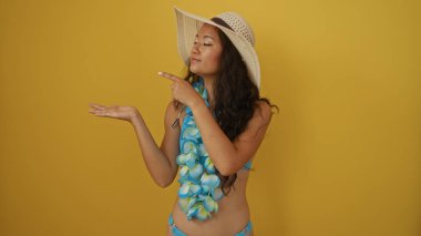 İzole edilmiş sarı arka plana karşı sunan güzel bir kadın kendine güvenen bir ifadeyle, çiçek çelengi ve hasır şapka takan yaz tatili temaları ve promosyon içeriği için ideal bir kadın.