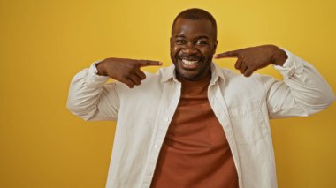 Genç adam, neşeli bir ifade ve rahat bir tavır sergileyen, canlı sarı bir arka plana karşı güvenle durarak gülümsemesini işaret ediyor..