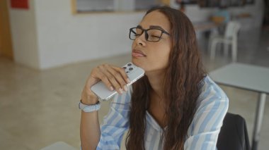 Gözlüklü genç bir kadın, iş yerinde, kapalı bir ofis ortamında akıllı bir telefon tutuyor.