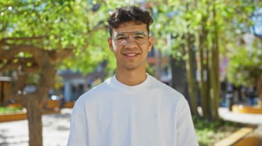 Beyaz gömlekli ve gözlüklü yakışıklı genç adam güneşli bir günde yeşil bir parkın içinde kendinden emin bir şekilde gülümsüyor..