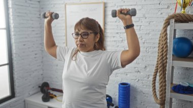 İspanyol orta yaşlı kadın spor salonunda egzersiz yapıyor. Parlak ve modern bir spor salonunda dambılları kaldırıyor..