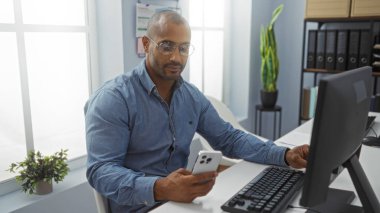 Ofiste çalışan yakışıklı, Afro-Amerikalı bir adam, modern bir çalışma alanında bilgisayarla bir masada otururken akıllı telefon kullanıyor..