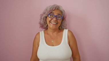 Gri saçlı, mavi gözlüklü, pembe izole edilmiş arka planda gülümseyen güzel, olgun İspanyol kadın..
