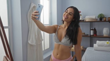 Spa merkezinde selfie çeken, iç çamaşırı giyen, esmer saçlı, gülümseyen ve kendine güvenen bir kadın..