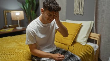 Genç bir İspanyol bir adam yatak odasında sarı bir yatak odasında oturuyor, bir eliyle gözünü ovuyor ve diğer eliyle gözlük tutuyor..