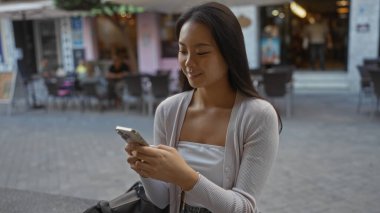 Asyalı bir kadın, arka planında kafeler olan bir caddede akıllı telefon kullanıyor, Çin 'de şehir dışı yaşam tarzını sergiliyor..