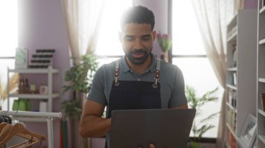 Dekoratif eşyalar ve bitkilerle çevrili, dizüstü bilgisayarı olan bir dükkânda çalışan genç adam..