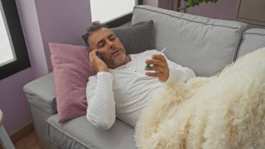 Yaşlı, İspanyol bir adam, elinde termometreyle bir kanepede dinleniyor ve yanında yastıkla rahat bir battaniyeye sarılıp telefonla konuşuyor..