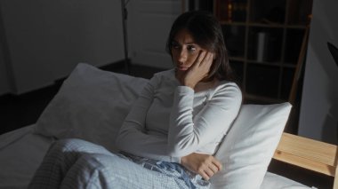 İspanyol esmer kadın, evinin loş ışıklı yatak odasında düşünceli bir şekilde yatakta oturuyor, rahat pijamalar giyiyor ve beyaz bir yastığa yaslanıyor..