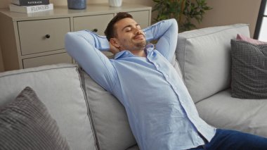 Sakallı yakışıklı, açık mavi tişörtlü, modern bir dairede, rahat, iyi dekore edilmiş bir oturma odasında, gri bir koltukta dinlenen bir İspanyol..