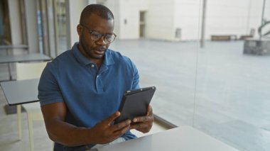 Mavi gömlekli genç bir adam ofiste tablet kullanıyor, işine odaklanmış ve meşgul, büyük bir pencerenin yanındaki masada tek başına oturuyor.