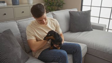 Genç bir İspanyol adam, rahat bir oturma odasında oturuyor, dachshund köpeğini tutuyor ve okşuyor. Bu arada ikisi de rahat, gri bir kanepede dinleniyor..