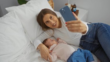 Kadın ve kızı rahat bir yatakta selfie çekiyorlar. Huzurlu bir yatak odası ortamında iç açıcı bir aile anını yakalıyorlar. Evdeki sevgi ve birlikteliği sembolize ediyorlar..