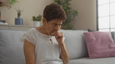 Kısa saçlı, yaşlı bir İspanyol kadın oturma odasında, üzerinde çiçekler ve yastıklar olan bir kanepede oturuyor. Düşünceli ve düşünceli görünüyor..