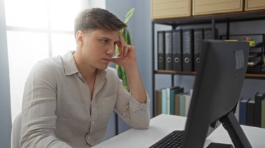 Modern ofis ortamında bilgisayarda çalışan genç adam, odaklanmış ve düşünceli görünüyor, arkaplanda klasörler ve dosyalar var.