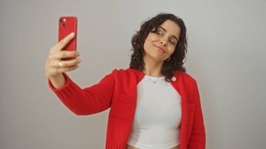 Genç, güzel bir İspanyol kadın kırmızı akıllı telefonuyla izole edilmiş beyaz arka plan duvarına selfie çekiyor..