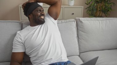 Sakallı, yakışıklı, Afro-Amerikalı bir adam mobilyalı bir oturma odasında rahatça oturuyor, gülümsüyor ve rahatlamış görünüyor..