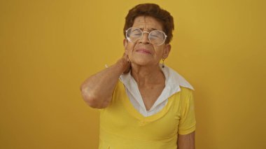 Sarı elbiseli, kısa saçlı ve gözlüklü yaşlı İspanyol kadın, izole edilmiş sarı arka planda duruyor, yansıtıcı görünüyor ve boynuna dokunuyor..