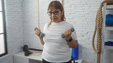 Orta yaşlı İspanyol bir kadın spor salonunda dambıllarıyla egzersiz yapıyor. İyi donanımlı bir spor merkezinde forma ve güç gösterisi yapıyor..