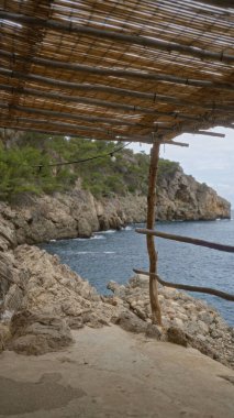 Mallorca, İspanya 'daki kayalık uçurumlara ve mavi Cala deia sularına bakan kırsal ahşap bir pavyonun güzel kıyı manzarası yemyeşil ve berrak gökyüzü ile çevrili..