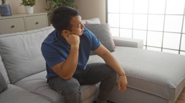 Genç Çinli bir adam modern bir oturma odasında oturuyor, düşünceli ve rahat görünüyor, mavi bir gömlek giyiyor..