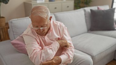 Bir kanepede oturup dirseğine bakarken gözlüklü ve gri saçlı, yaşlı, beyaz bir adam..