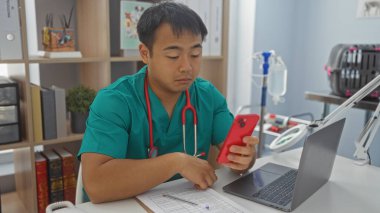 Veteriner kliniğinde çalışan genç Çinli bir adam, ameliyat önlüğü ve steteskop takıyor, elinde belgelerle masada otururken akıllı telefon kullanıyor..