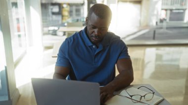 Genç, Afro-Amerikalı bir adam ofis ortamında dizüstü bilgisayarla çalışıyor. Masasında gözlük ve not defteriyle dikkatle çalışıyor.
