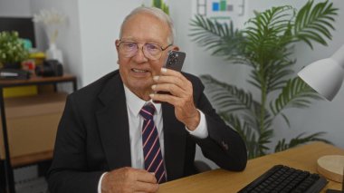 Takım elbiseli, elinde akıllı bir telefon olan, ofisteki bir masada oturan, arka planda iç mekan bitkileri ve ofis ekipmanlarıyla kameraya bakan yaşlı beyaz bir adam..