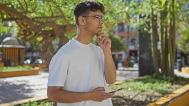 Şehir parkında elinde akıllı bir telefonla dikilen genç bir İspanyol, şehir merkezinde düşünceli görünüyor..