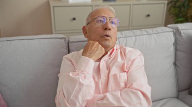 Gri saçlı, gözlüklü, pembe tişörtlü, gri bir kanepede düşünceli, rahat bir oturma odasında oturan yaşlı beyaz bir adam..