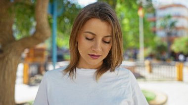 Çekici sarışın kadın gün boyunca şehir parkında duruyor, sakin bir ifadeyle, arka planda ağaçlar ve şehir unsurlarıyla çevrili..