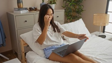 Esmer saçlı genç bir kadın yatak odasındaki bir yatakta oturmuş dizüstü bilgisayarını kullanırken gözlüklerini ayarlıyor, etrafı dekorasyon malzemeleriyle çevrili..