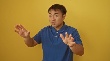 Mavi gömlekli genç Asyalı adam, izole edilmiş sarı arka plana karşı jest yapıyor..