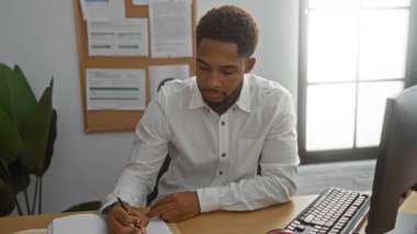Ofiste çalışan genç bir adam, elinde kalemle bir kağıda yazı yazıyor, arka planda bir ilan panosuyla bilgisayarın önünde oturuyor..