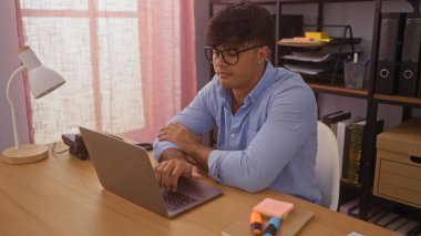 Modern ofis ortamında dizüstü bilgisayarda çalışan yakışıklı İspanyol bir adam. Etrafı kitaplarla ve kırtasiyelerle çevrili. Üretken bir çalışma ortamını simgeliyor..