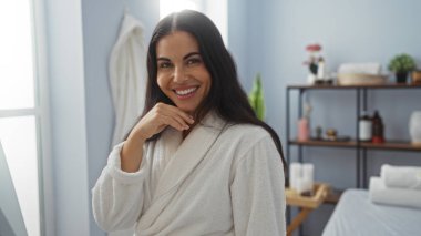 Sağlık merkezinde beyaz bornoz giymiş, kaplıca odasında gülümseyen bir kadın.