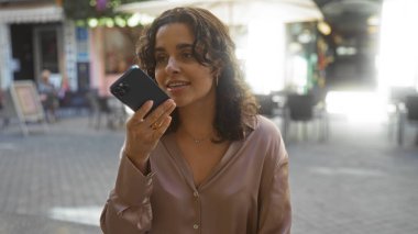 Esmer saçlı, genç, çekici bir İspanyol kadın canlı bir sokak ortamında akıllı bir telefonla konuşuyor. Kasabanın canlılığını yakalıyor..